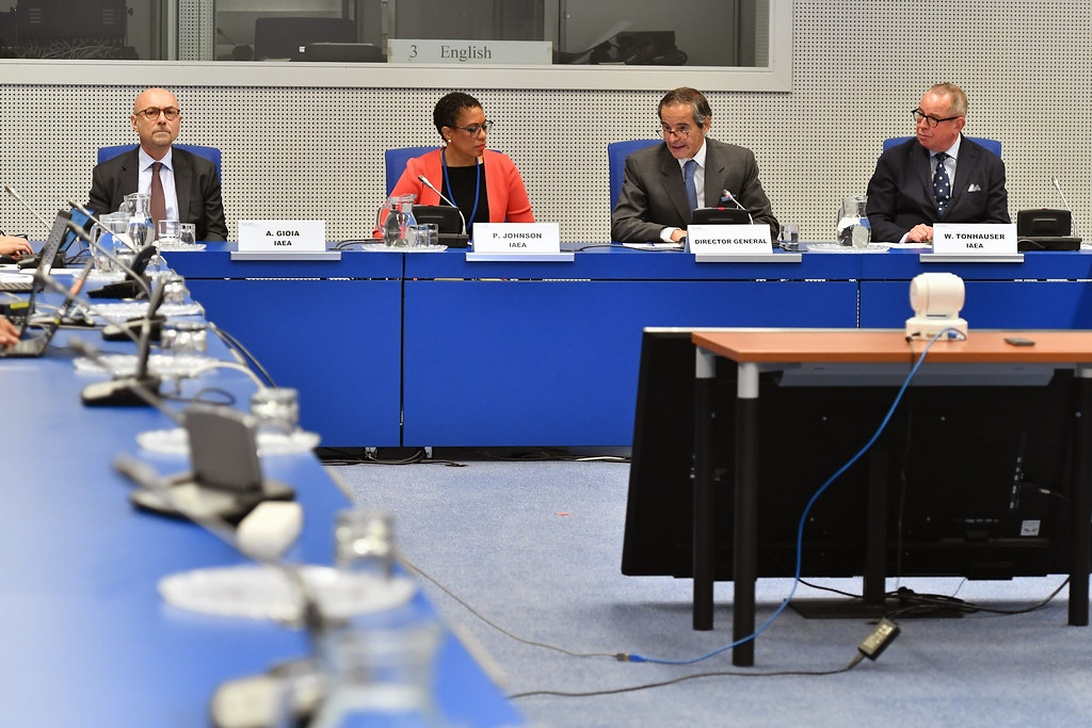 Delegaten tijdens een formele conferentie van een internationale organisatie, mogelijk de IAEA, aan een blauwe tafel met microfoons.