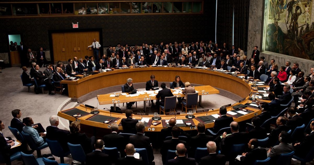 Een formele, ronde vergaderzaal waar delegaten rond een grote tafel zitten, lijkend op de VN Veiligheidsraad.